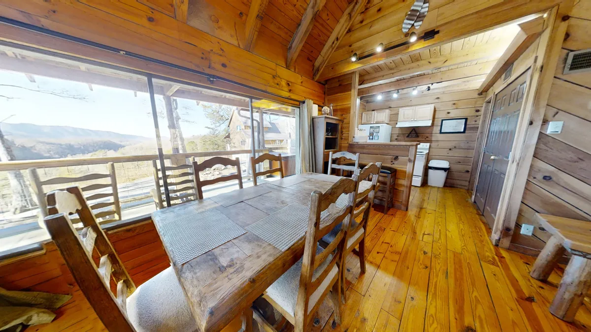 This image showcases the dining table set near expansive windows, exposed wooden beams overhead, and an adjacent kitchen area. The setup is ideal for meals, games, and conversations, depicting an inviting, communal space flooded with natural light.