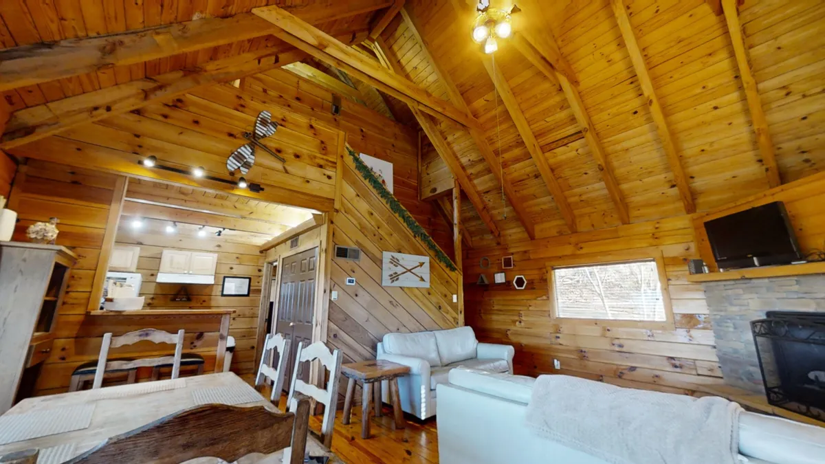 This image captures a broader view of the living/dining area, featuring wooden ceiling beams, a stone fireplace, comfortable seating, and a dining table. The space feels open and welcoming, designed for group activities like sharing stories or enjoying meals together.