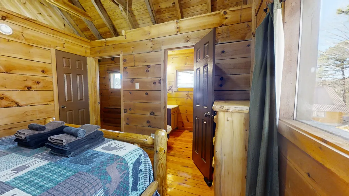 This image shows a warmly lit bedroom featuring a rustic log bed, towels neatly placed on the bed, and an open door leading to the bathroom. The space is framed by wooden walls and allows in natural light, creating a cozy and inviting atmosphere that suggests moments of gathering and relaxation after outdoor adventures.