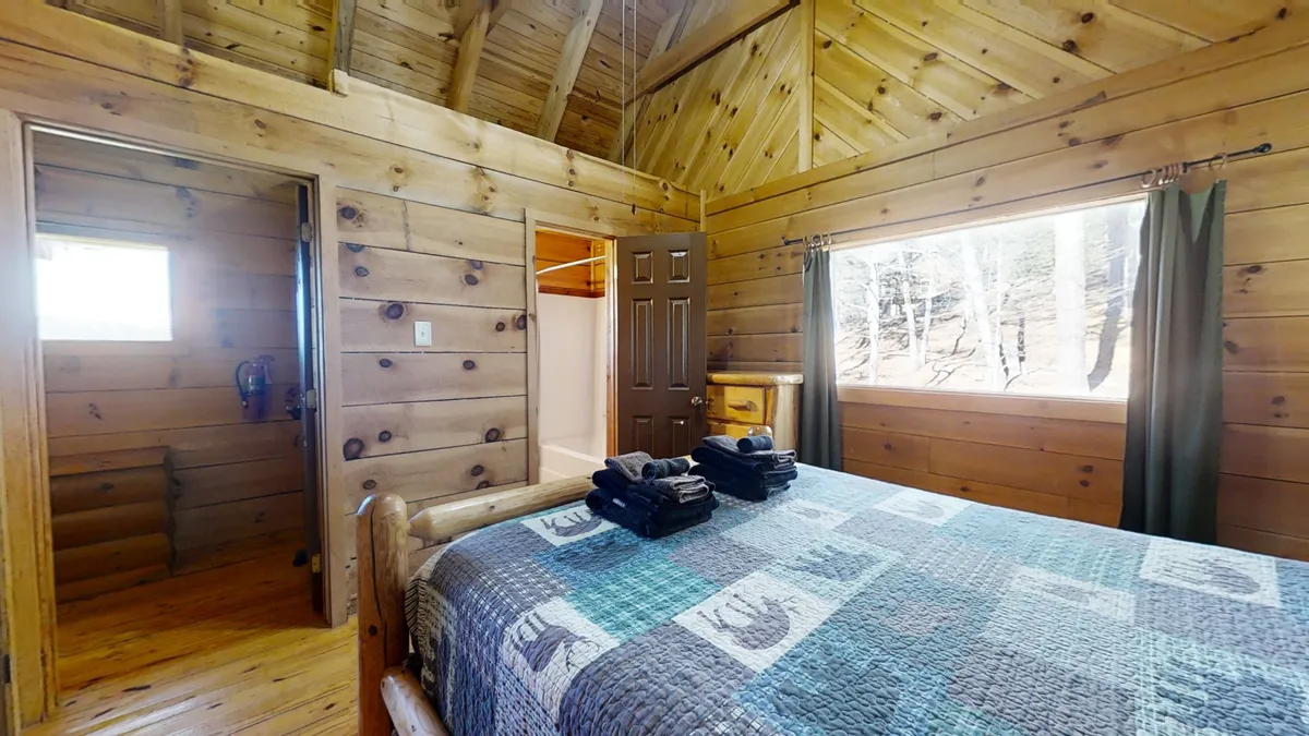 This tranquil bedroom features a wooden bed frame adorned with a patchwork quilt, a large window that fills the space with natural light, and warm wooden paneling throughout. The scene is peaceful, with a focus on relaxing, reading, or simply enjoying the views of nature from inside the cabin.