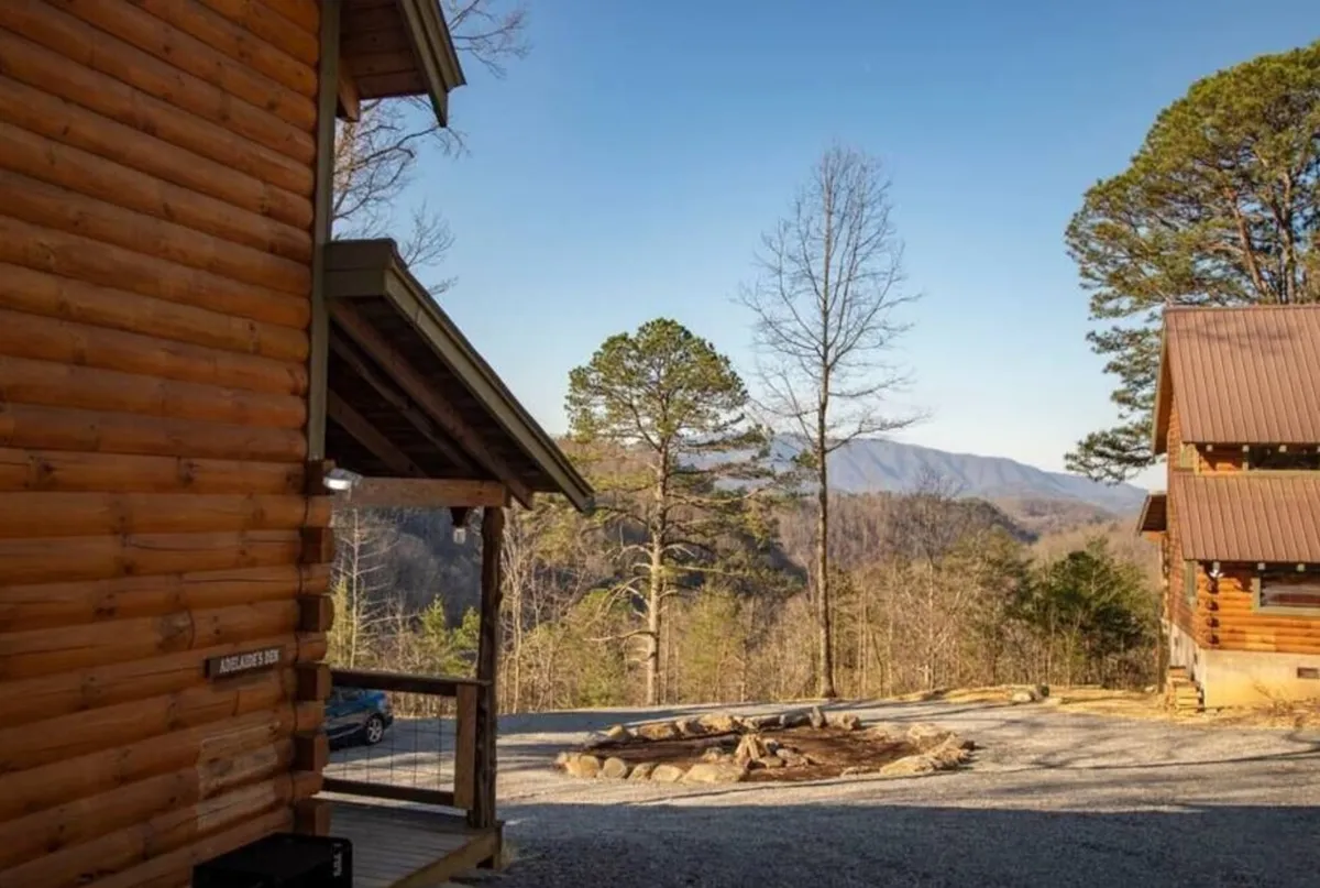 This exterior shot shows the log cabin set against a stunning mountain backdrop. The fire pit area is visible in the yard, surrounded by trees and ample outdoor space. The scene captures a tranquil, inviting atmosphere ideal for gatherings around the fire, making s'mores, and stargazing under the Tennessee sky.