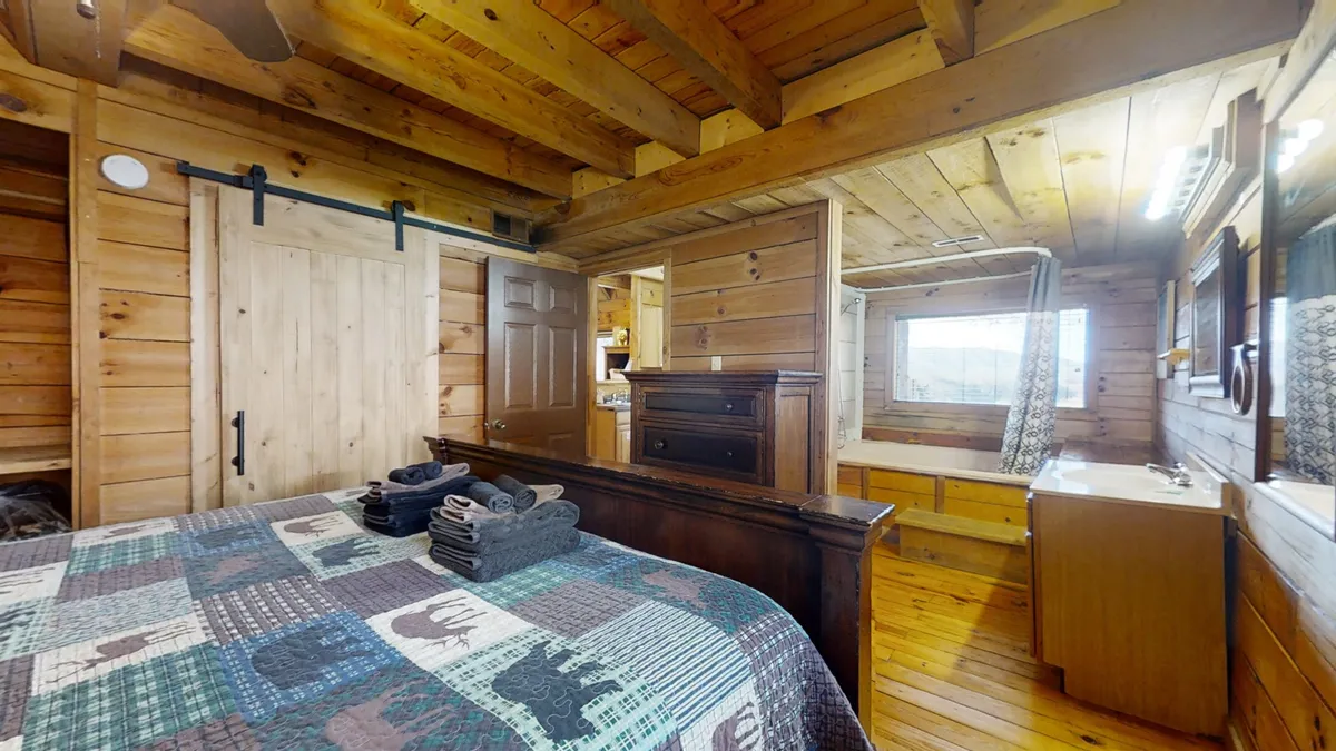 A comfortable bedroom with wooden beams, a bed adorned with a patterned quilt, and a vanity area with a mirror. Soft morning light pours in from a window, creating a warm and inviting ambiance for relaxation and privacy.