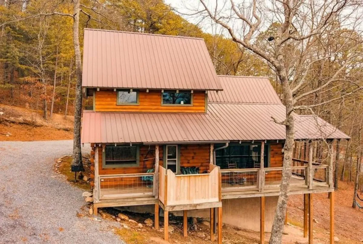 This image features a charming wooden cabin with a metal roof tucked among lush trees. The covered deck is visible, furnished with rocking chairs, inviting guests to relax and take in the panoramic mountain views. The scene suggests a place for sunrise coffees or sunset unwinding, and hints at the outdoor lifestyle the section describes.