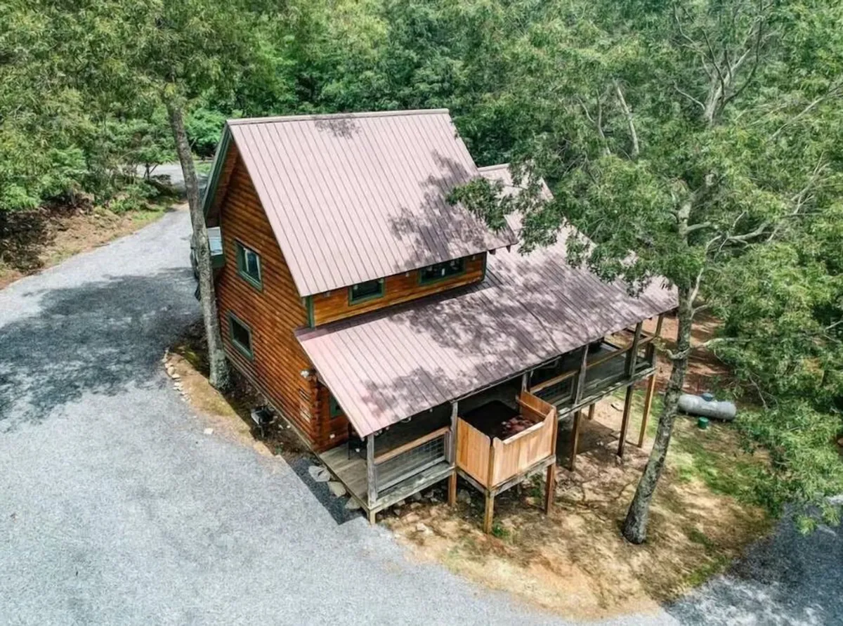 This exterior image captures the inviting wooden cabin surrounded by lush trees, with a gravel driveway leading up to it. The cabin features a metal roof and a spacious porch, all set in a peaceful mountain environment. The view hints at the tranquility and natural beauty that families can enjoy during their stay, serving as a perfect base for adventures in the Smoky Mountains.