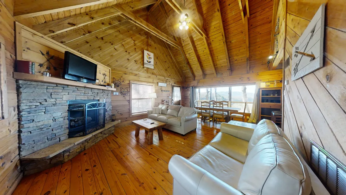 The image showcases a warmly lit living room with a prominent stone fireplace, a large window offering views of the outdoors, a comfortable leather sofa, and a wooden dining table. The overall setting exudes warmth and comfort, perfect for family gatherings and relaxation. The stone fireplace and leather seating align perfectly with the section's features.