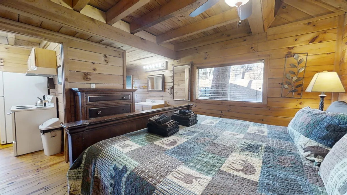 An inviting suite with a comfortable bed covered in a plush quilt, wooden furniture, a small kitchenette, and large windows letting in natural light. The space is both functional and cozy, ideal for longer stays or family needs.
